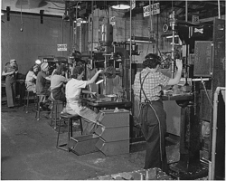 Una foto de la época de la II Guerra Mundial de cinco mujeres trabajando en taladros, todas vestidas con pantalones, camisas de trabajo, sobreros y mandiles.