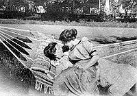 Foto en blanco y negro de dos mujeres sentadas en una hamaca, vestidas con la moda de principios del siglo XX; una está reclinada y la otra está sentada a su cintura y la abraza, ambas mirándose.
